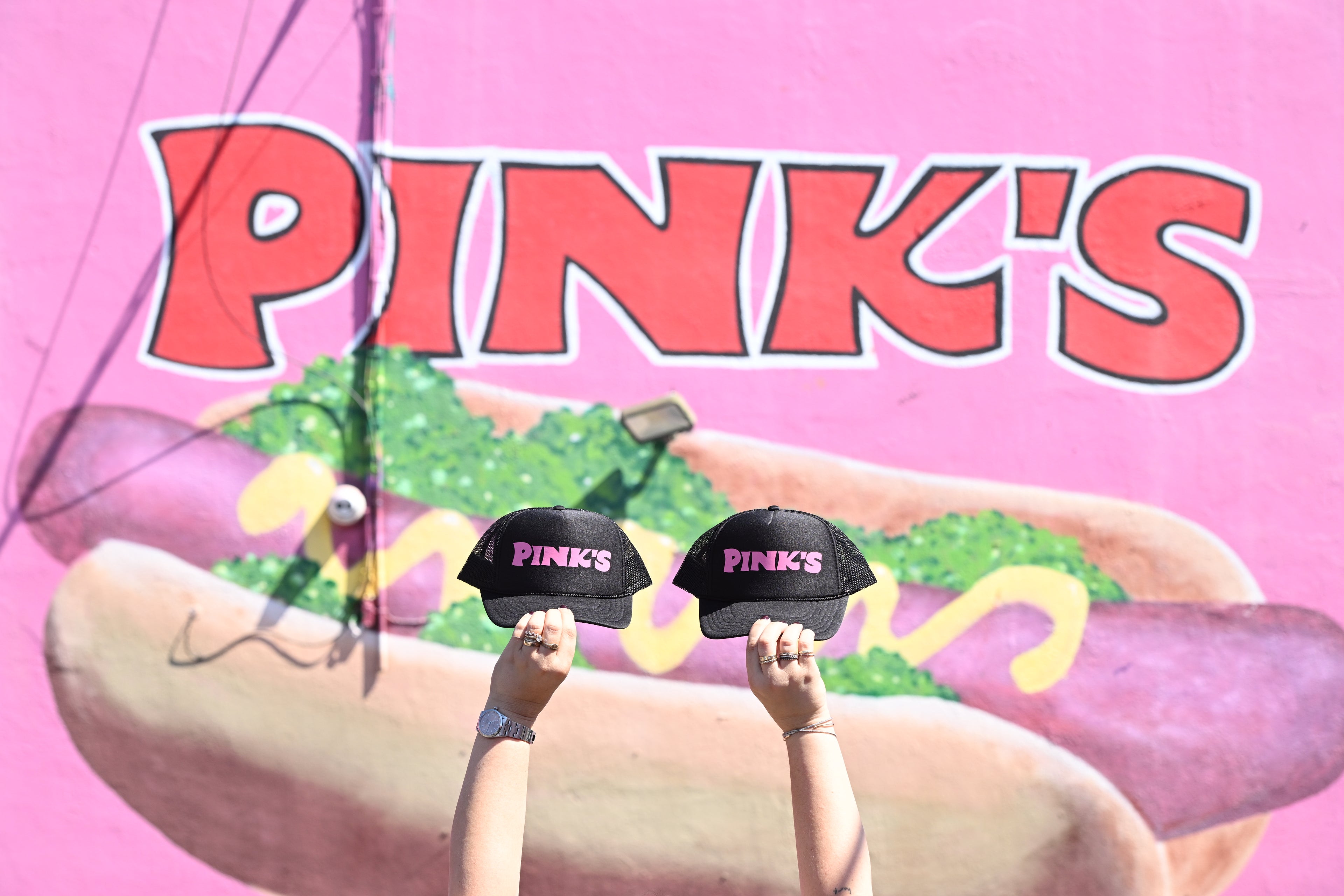 Hands holding up two of the new Pink's logo trucker hats